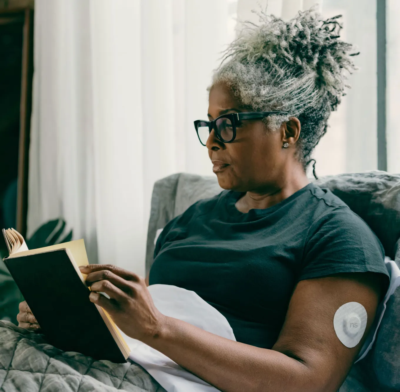 A Nutrisense member reading a book on bed while wearing a CGM sensor on her left upper hand