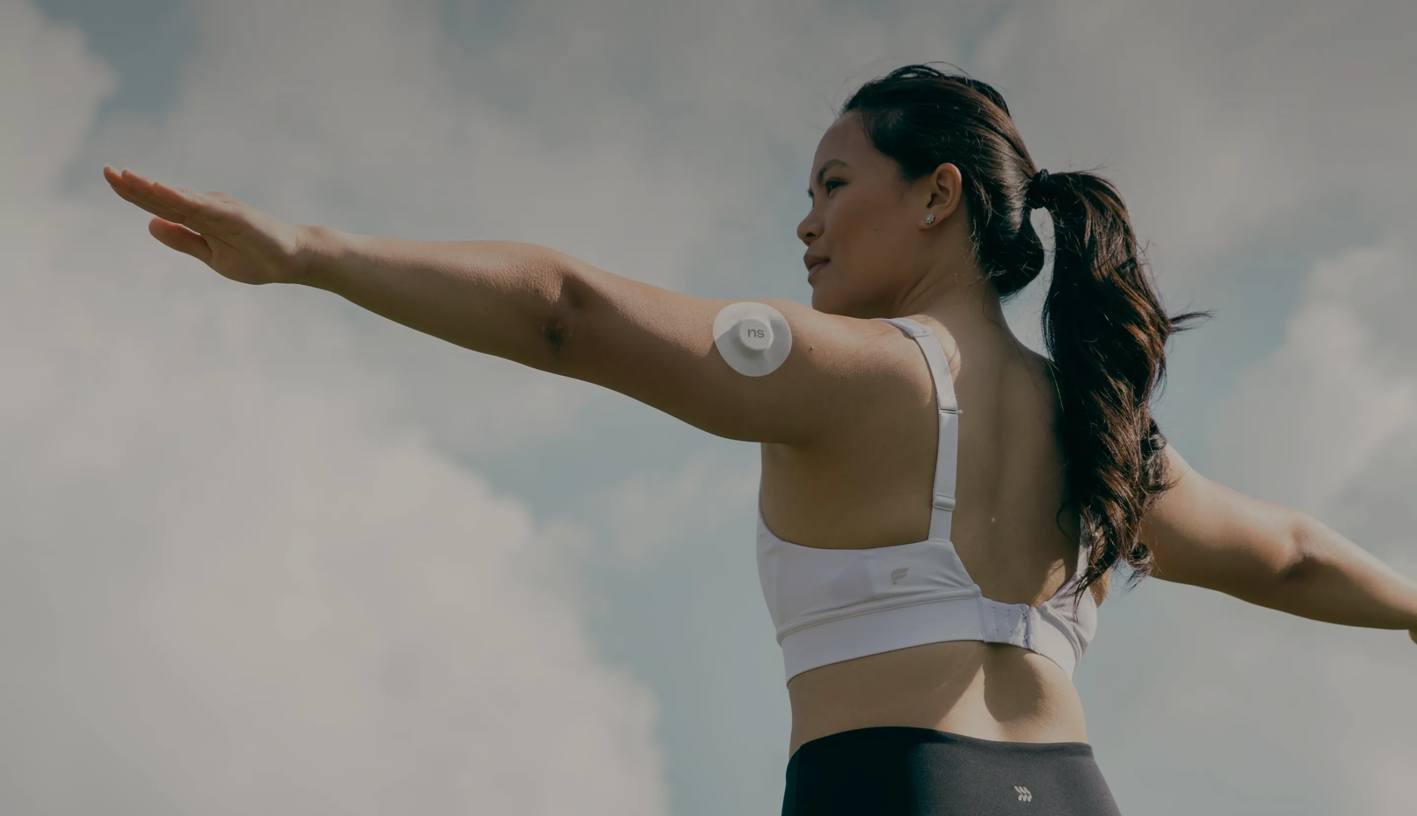 Back view of lady standing facing her left side while stretching her hands wide aside wearing Nutrisense CGM device on her left upper hand available at nutrisense.io