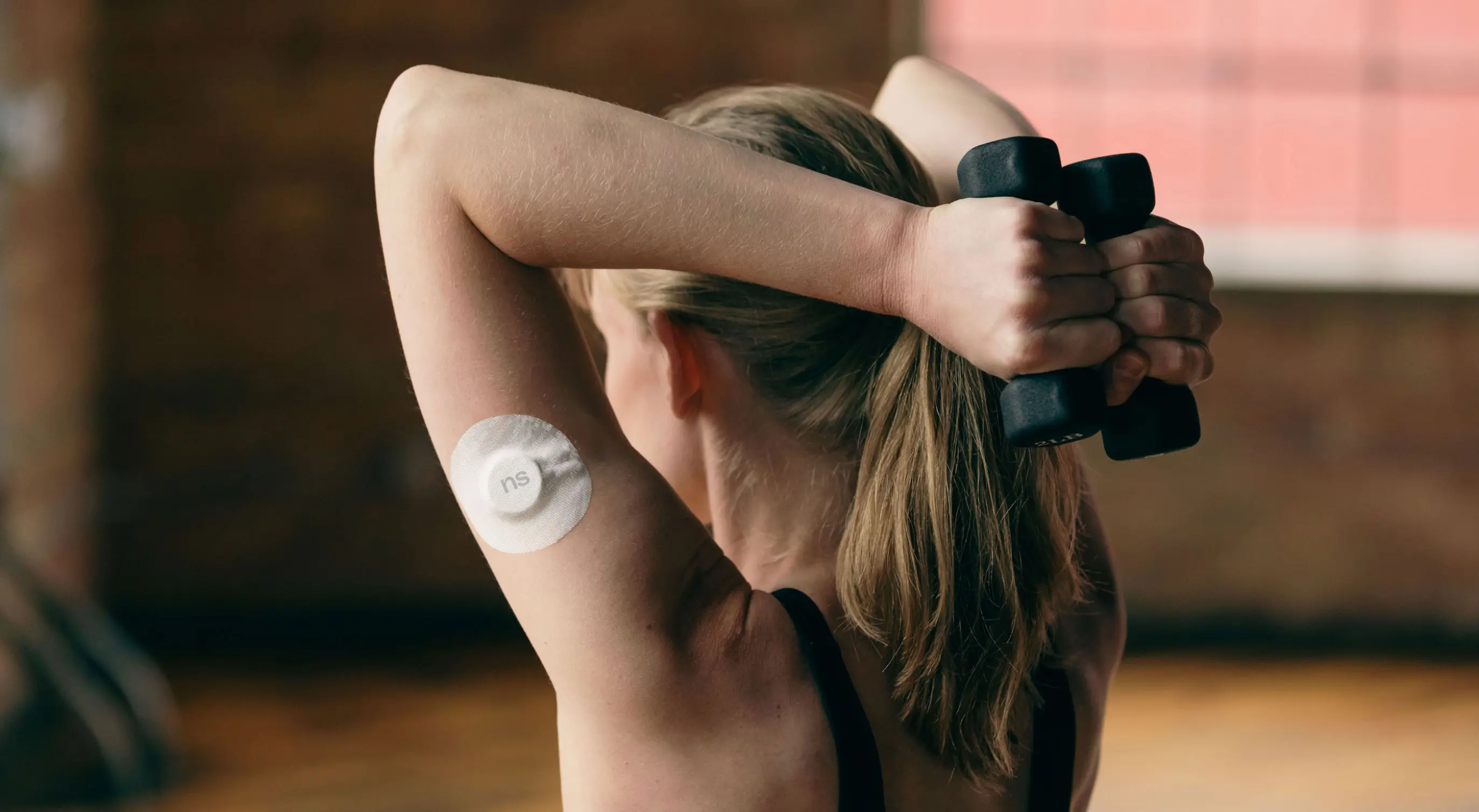 A Nutrisense member holding dumbbells behind her back with a Nutrisense cgm on her left upper hand