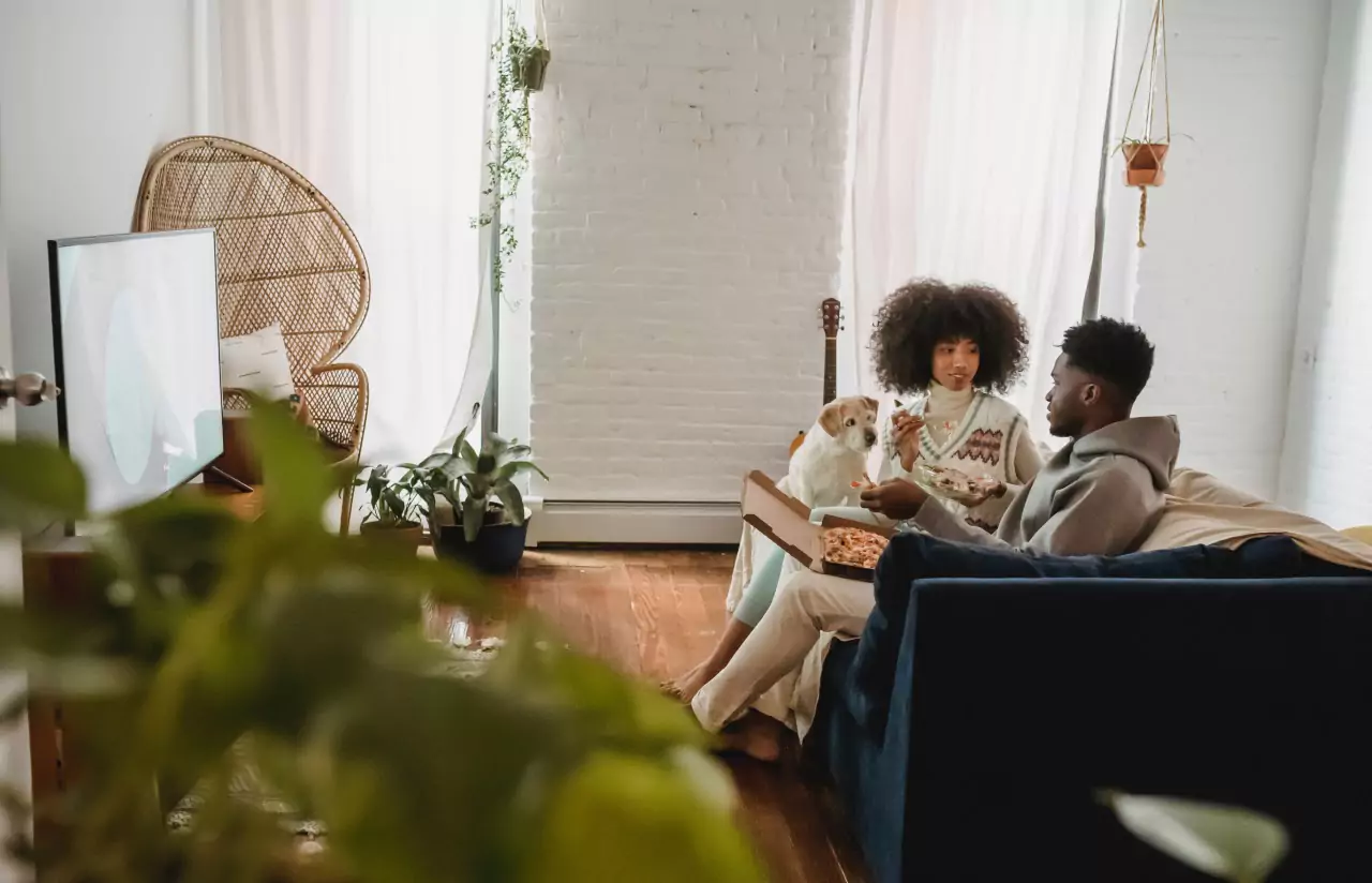 A woman and man sitting on  couch eating pizza, with a puppy watching them 