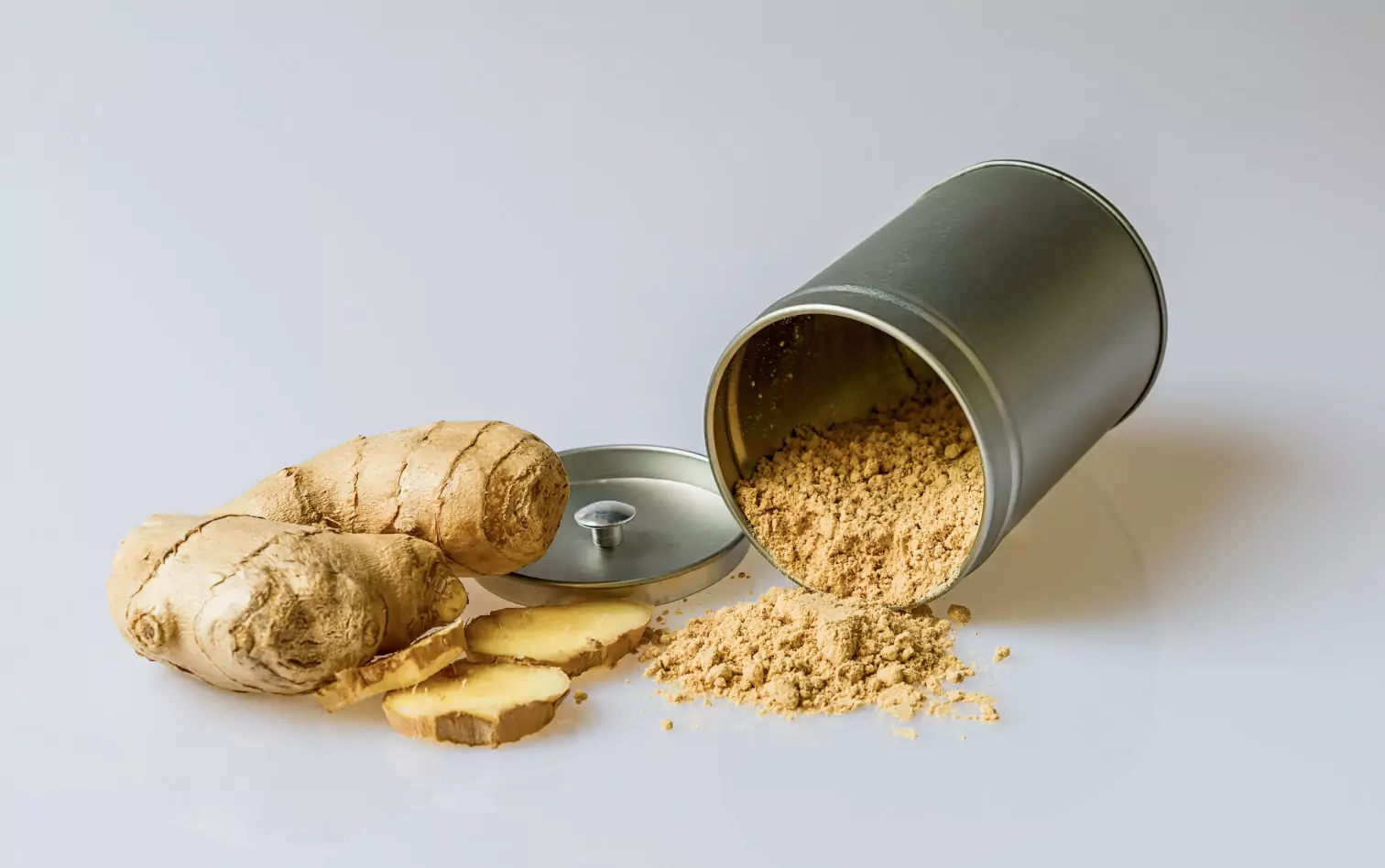 A steel canister lying sideways on a white backdrop, powdered ginger spilling out and fresh ginger on the left