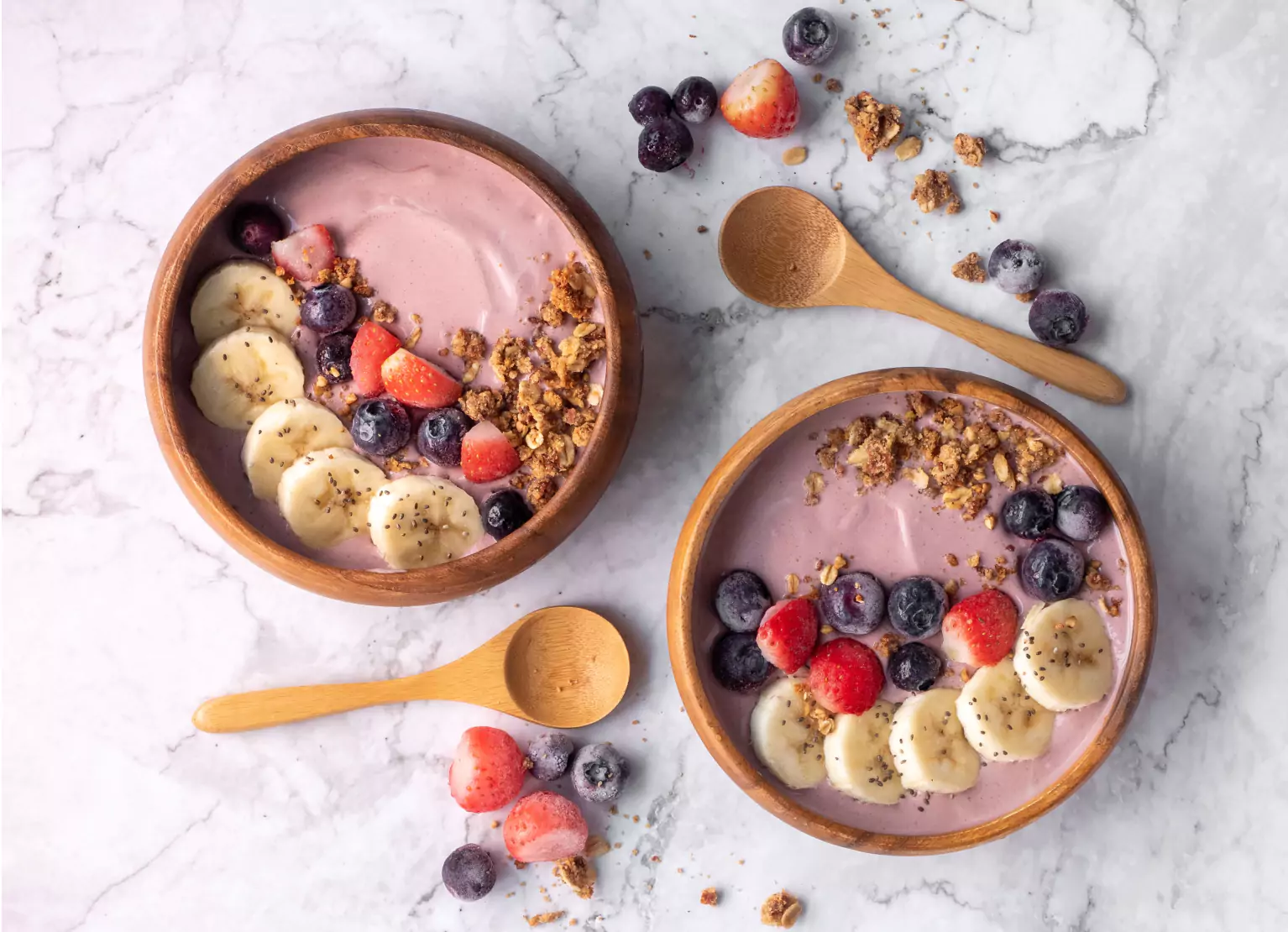 Two wooden bowls of strawberry yogurt bananas, strawberries, blueberries and granola topping. Two wooden spoons and berries alongside