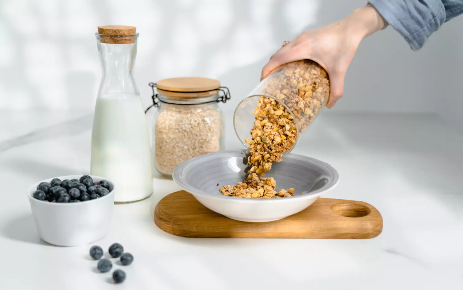 a person pouring granola into a bowl, a bowl of blueberries and a bottle of milk 