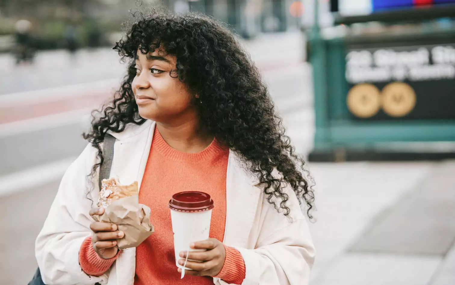 A woman crossing the street holding a sandwich and a cup of coffee to go