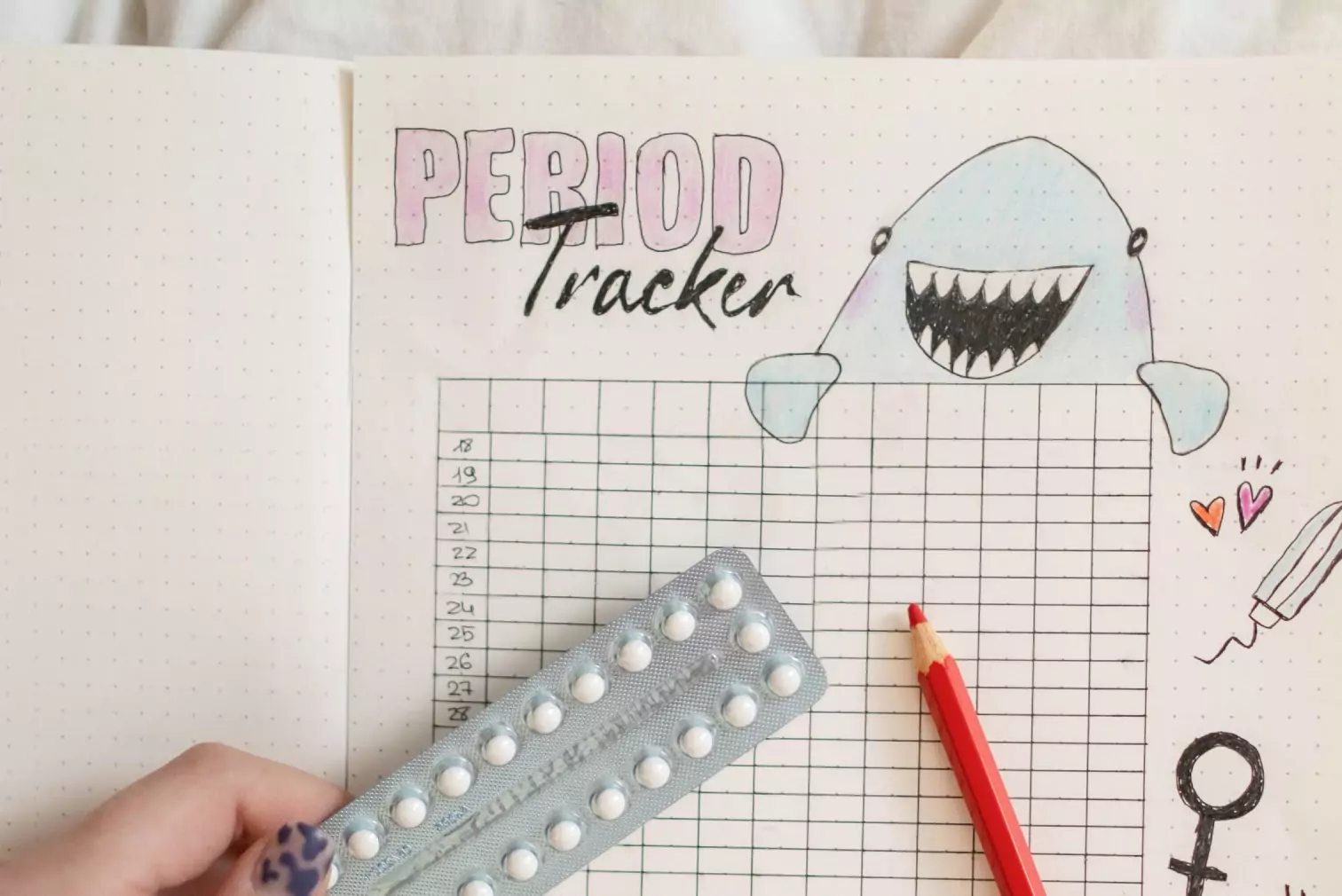 A book with an illustration of a period tracker, a picture of a shark. A color pencil and a pack of birth controls lie on top of the page 