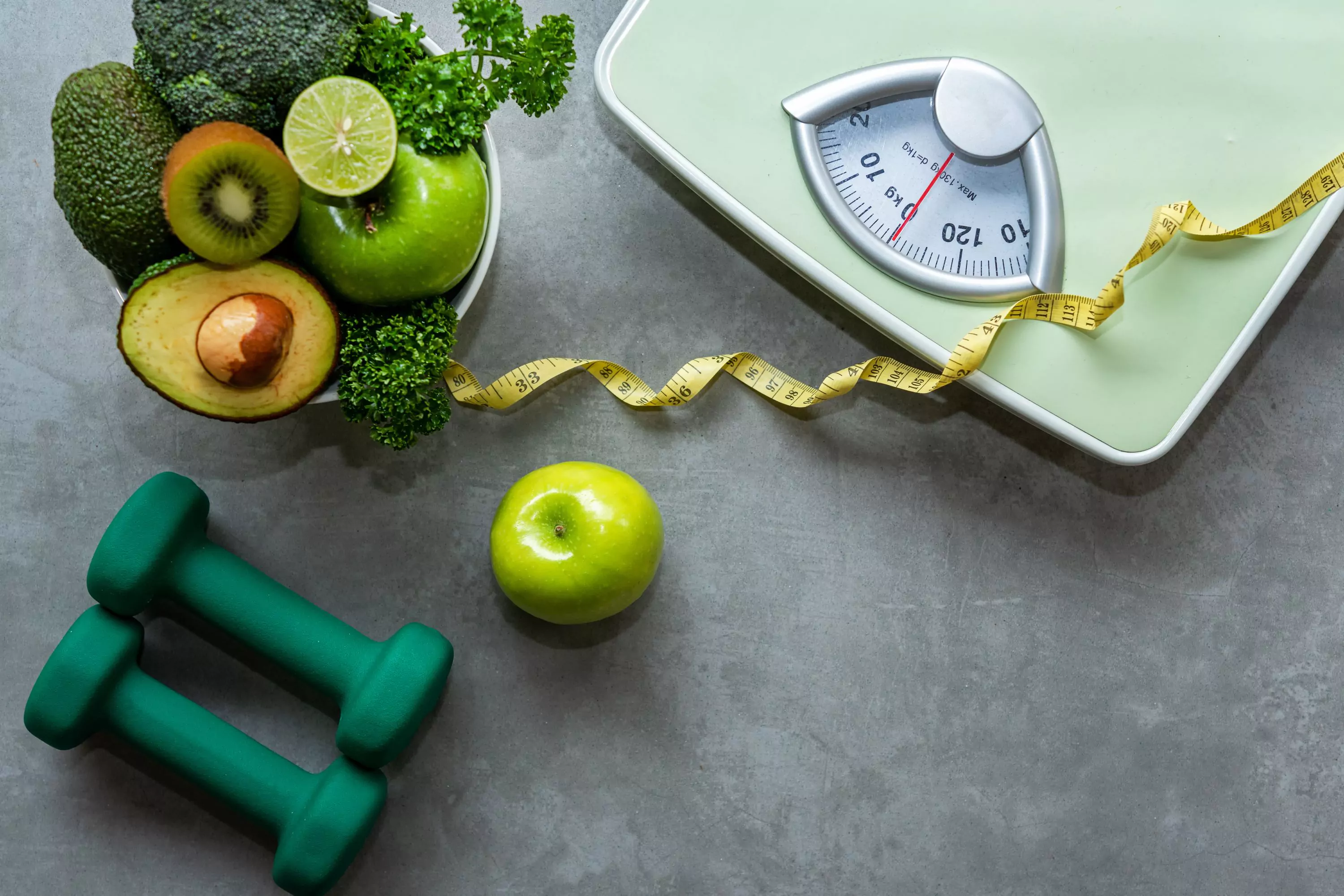 a scale, a tape measure, dumbbells and a bowl of green veggies and fruits