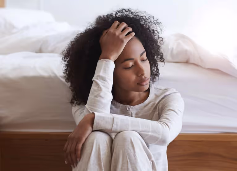 A woman in white pyjamas sitting at the foot of a bed holding her forehead