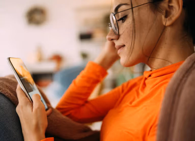 Side profile of a woman in an orange top, wearing glasses, looking into a martphone