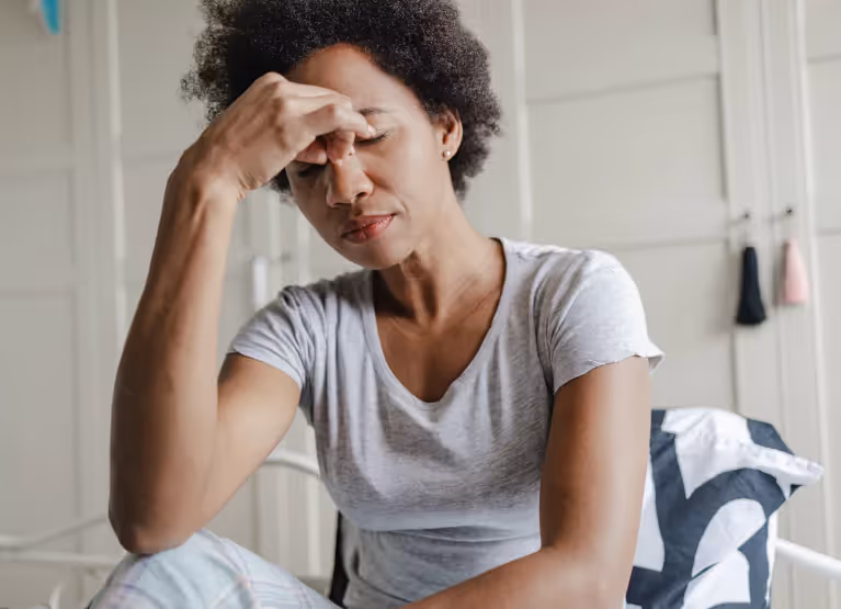 A woman in grey t shirt and slacks sitting in front of a white cupboard holding her forehead