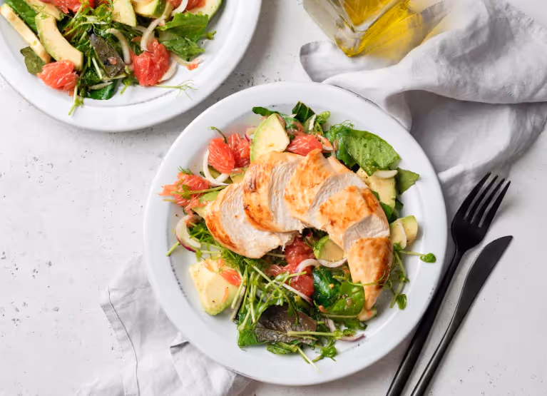 Two plates of leafy greens, avocado, grapefruit, and chicken breast