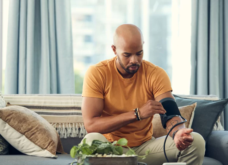 Man in orange T shirt taking his own blood pressure while sitting down on the couch