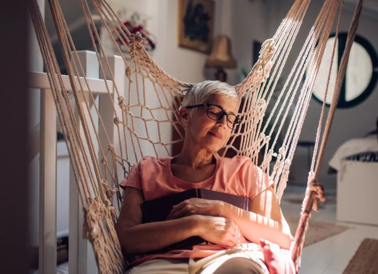 An older person in a pink tshirt with short grey hair and glasses, lying in a hammock swing