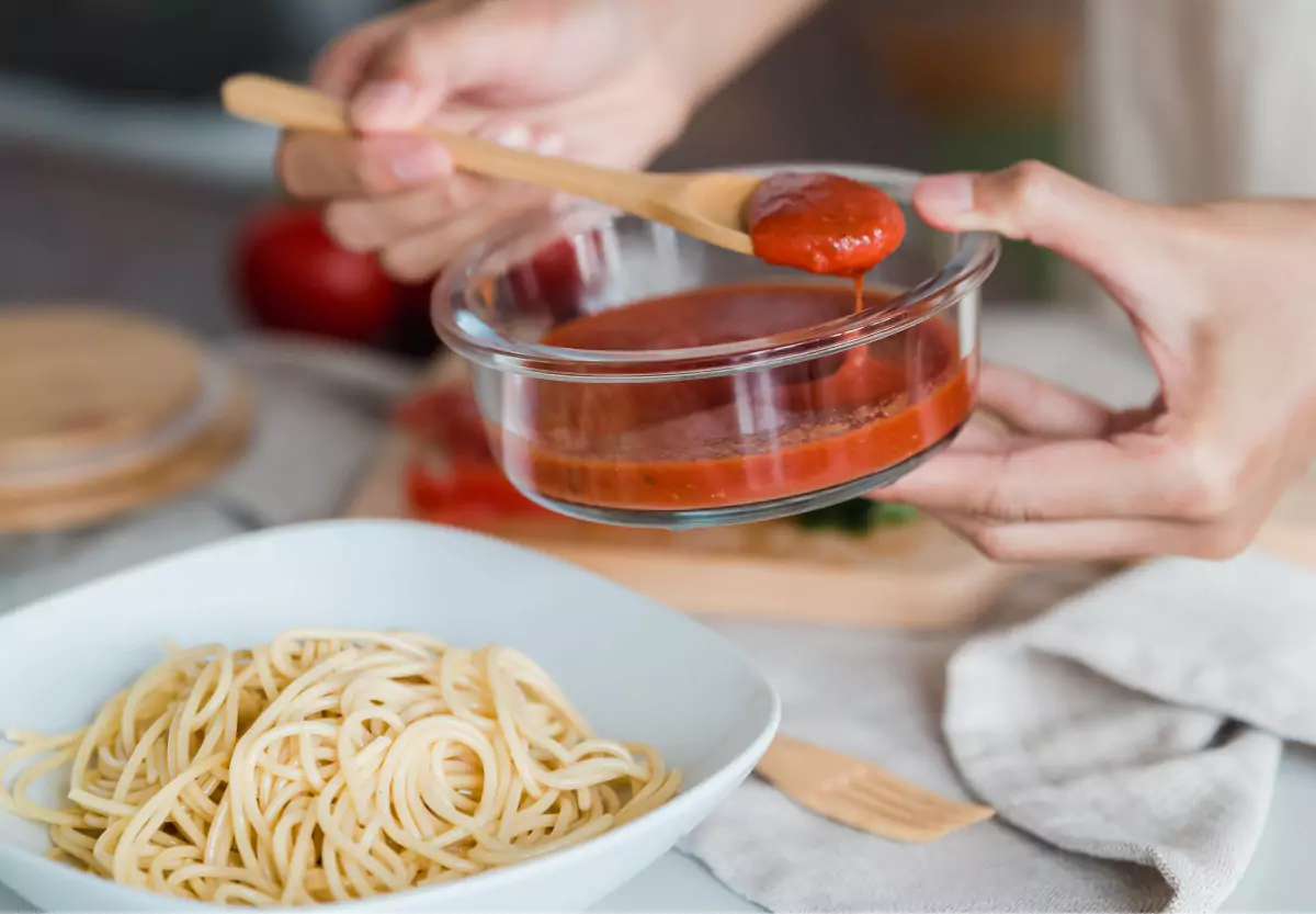 A closeup of hands holding a wooden spoon and a bowl of red pasta sauce