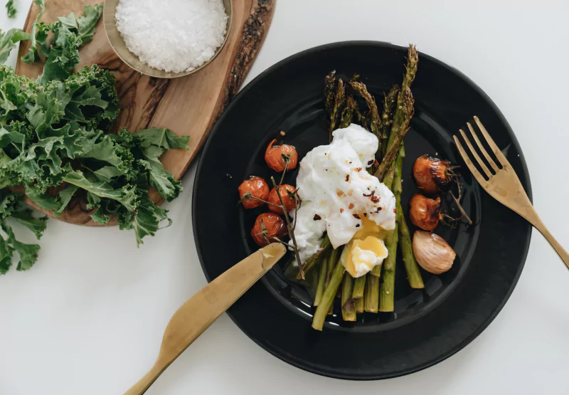 A black plate with a poached egg, tomatoes, asparagus and garlic with greens and salt in the background