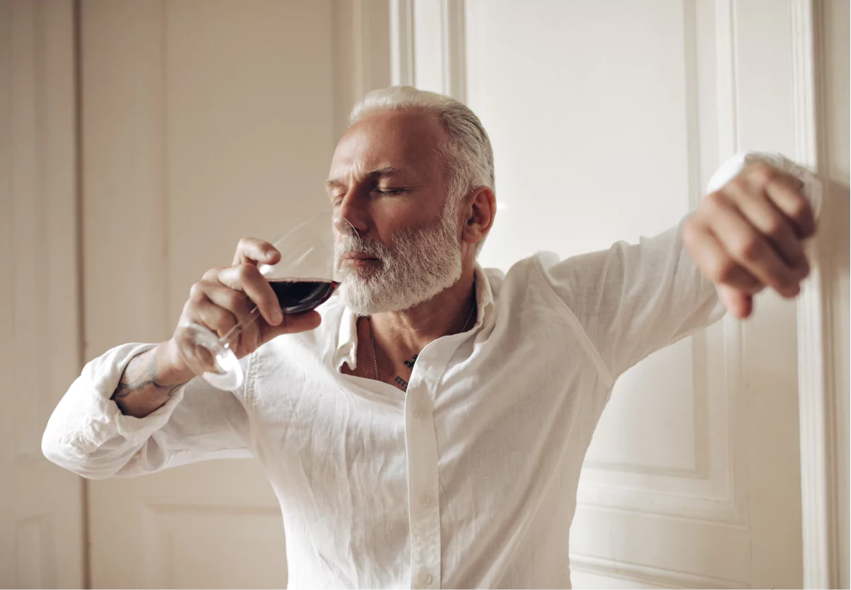 An older man in a white shirt leaning against a wall drinking a glass of red wine