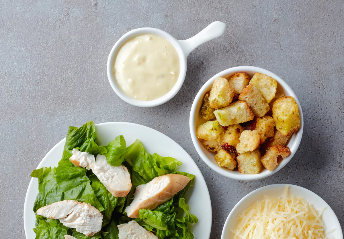 a bowl of greens with chicken, a bowl of croutons, and caesar dressing