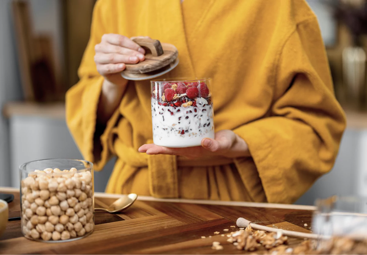 A person in a yellow robe, holding a glass canister of berries and yogurt in one hand and wooden cover in the other