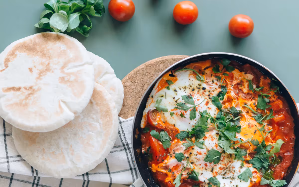 A bowl of Shakshouka lunch with pita bread and cherry tomatoes on the side, against a blue backdrop