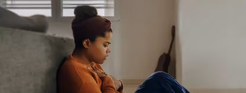 A woman sitting on the floor at the foot of a bed, holding her chest