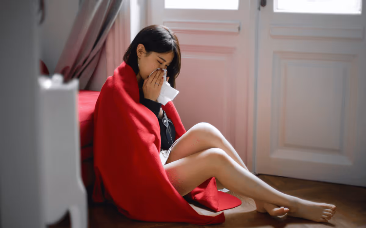 A woman with long dark hair sitting on the floor, wrapped in a red blanket and using a tissue