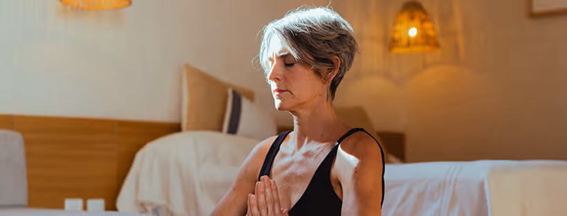 A person with short grey hair sitting on the floor by a bed, meditating