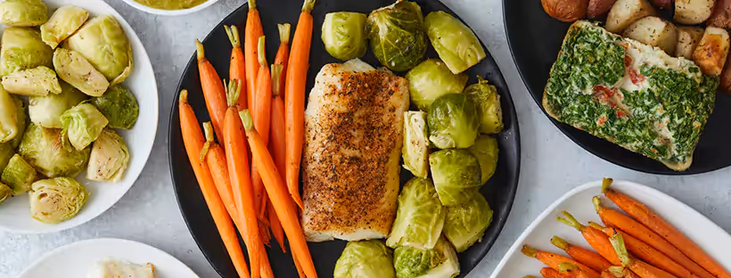 A close-up of a meal of protein, carrots and Brussels sprouts on a black plate, surrounded by other plates of similar food