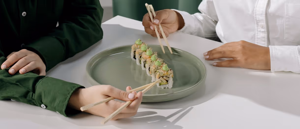 Two pairs of hands, one in a green shirt and one in white, sharing a plate of sushi rolls