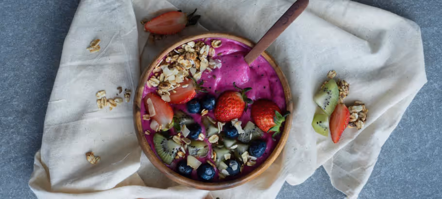An overhead view of a white bowl filled with the fruit, nuts, granola, yogurt and other contents of an Acai Bowl, placed on a white kitchen cloth