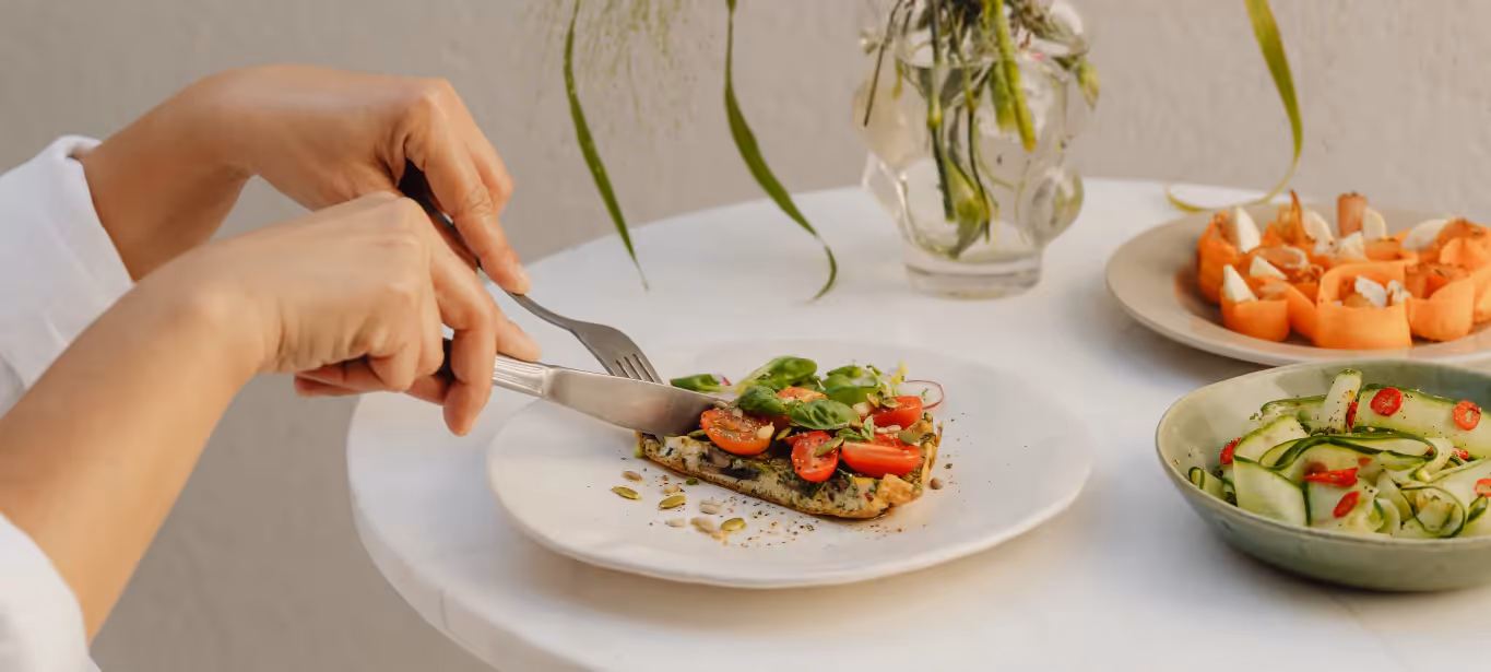 Hands holding cutlery, cutting into an open-face vegetable sandwich on a white plate