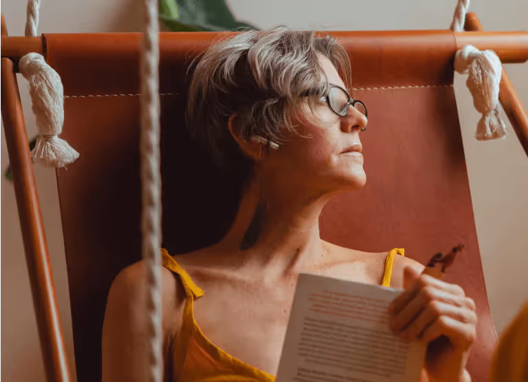 An older person with short grey hair, in glasses, sitting down reading a book and looking away pensively