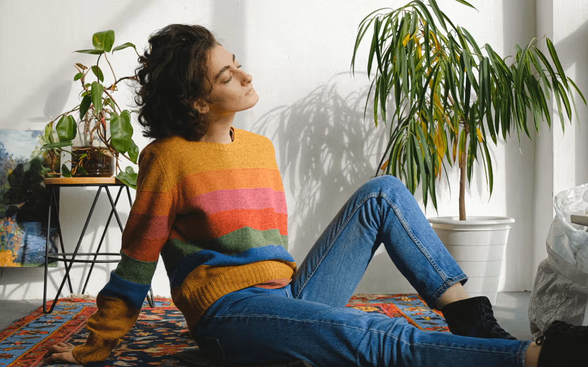 A person in a colourful striped sweater and blue jeans, with dark hair sitting on a carpet, with houseplants in the background