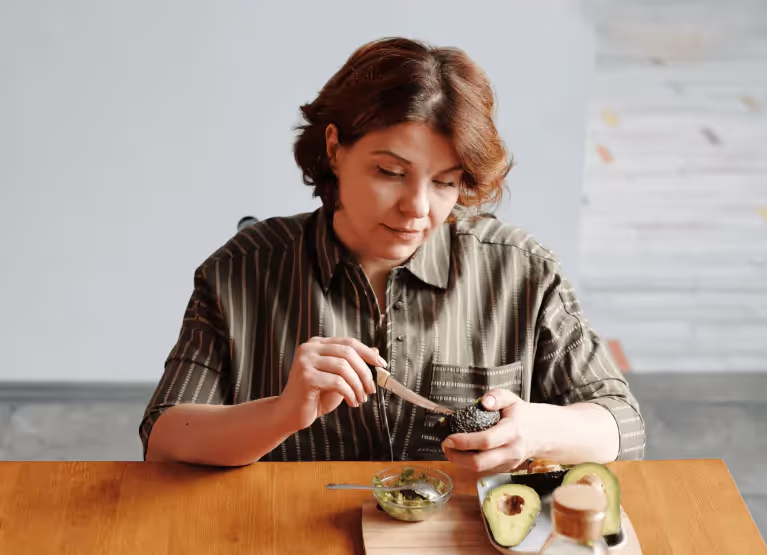A person with mid-length brown hair sitting at a wooden table eating an avocado