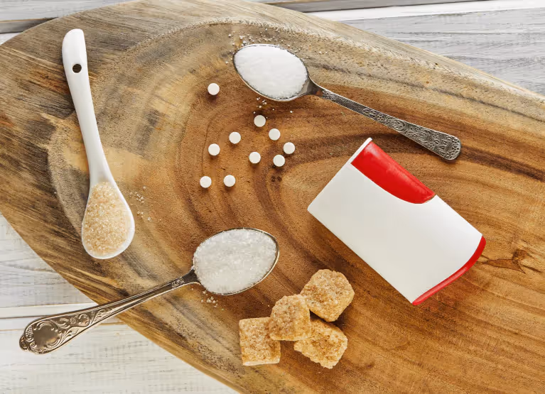 A wooden board with silver spoons filled with brown and white sugar, with brown sugar cubes on the side.
