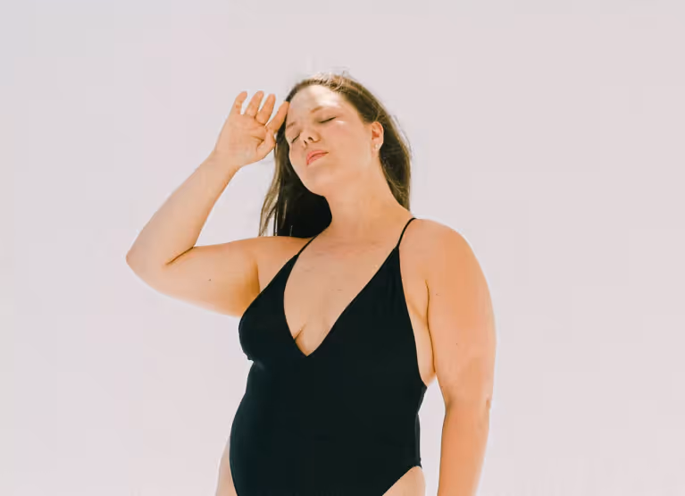 Someone in a black bathing suit with long, black hair, standing outdoors with their hand on thier forehead