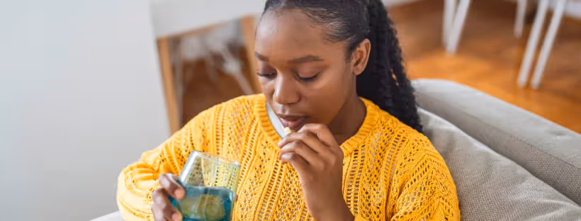 A woman in a yellow sweater sitting on a couch holding a blue glass of water in one hand and a pill in the other