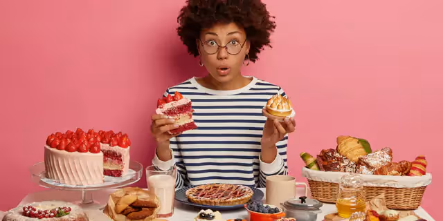 a person holding a piece of cake sitting at a table full of different kinds of sweets