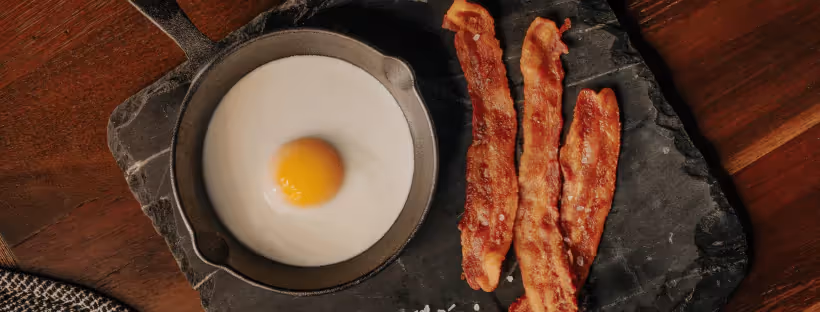 A cast iron pan with a sunny side up egg and two strips of fried bacon at the side