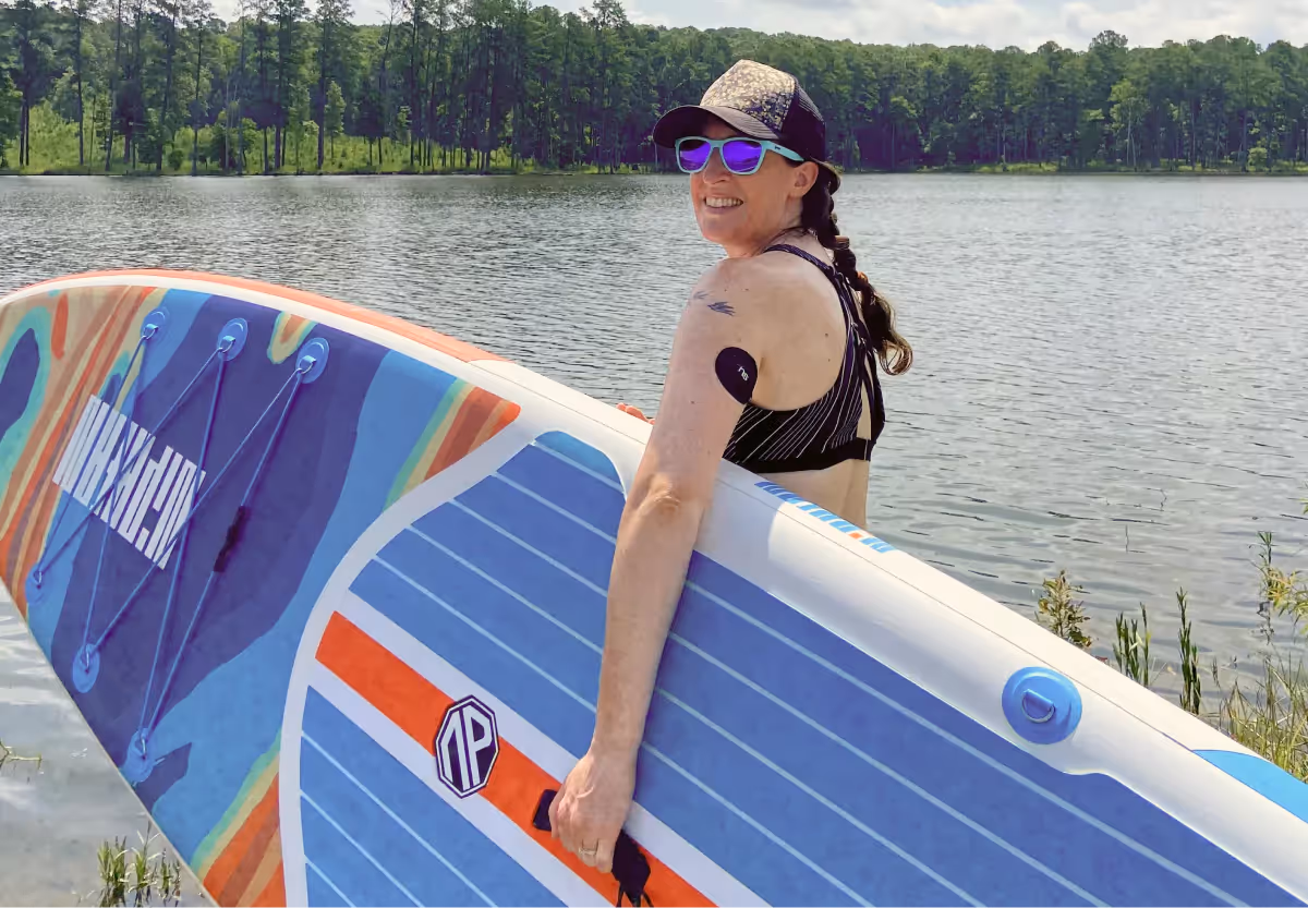 Bridget Fletcher holding a surfboard by a body of water outdoors wearing a cgm with a nutrisense branded patch