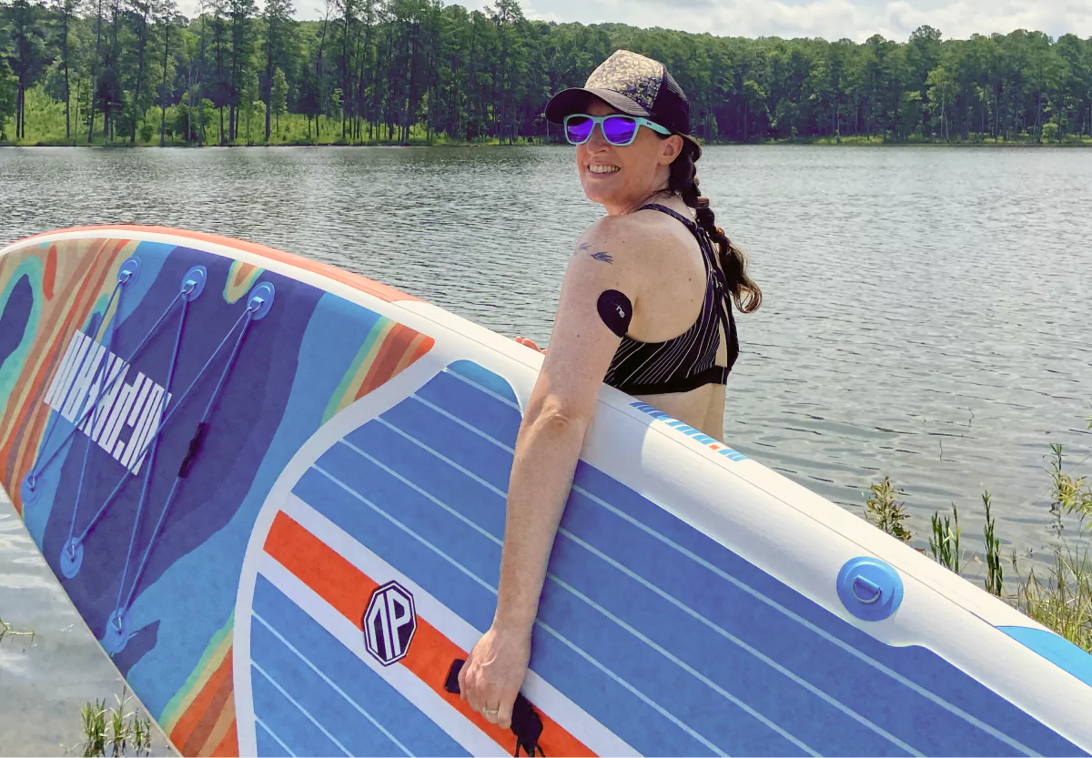 Bridget Fletcher holding a surfboard by a body of water outdoors wearing a cgm with a nutrisense branded patch