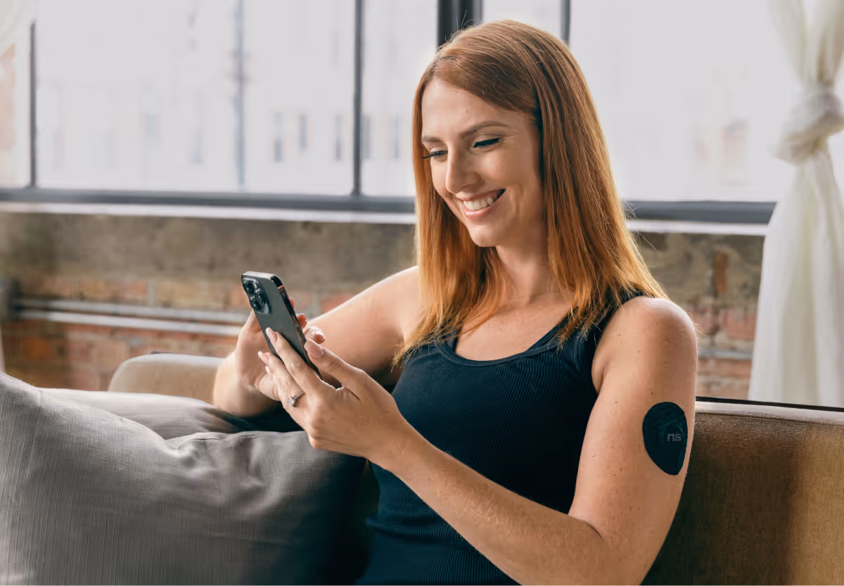 A person with red hair sitting on a couch looking at their mobile, wearing a cgms sensor with a nutrisense branded patch