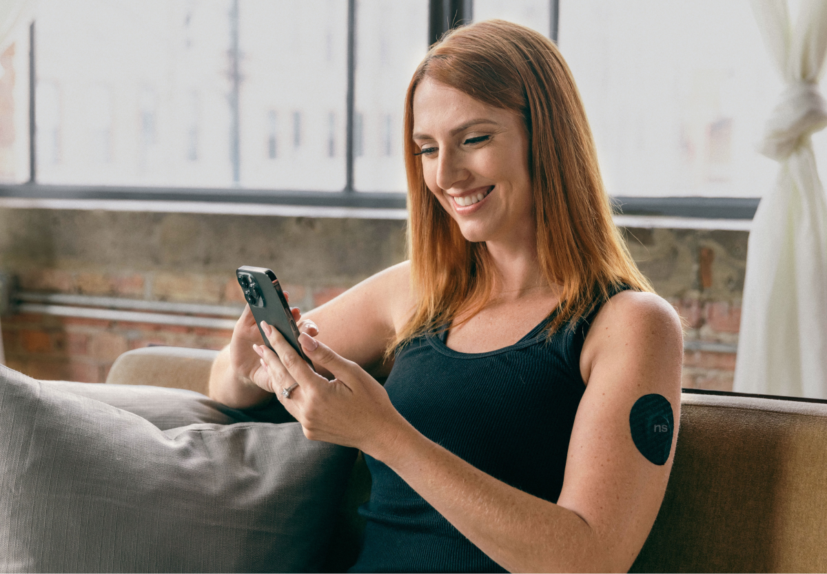 A person with red hair sitting on a couch looking at their mobile, wearing a cgms sensor with a nutrisense branded patch