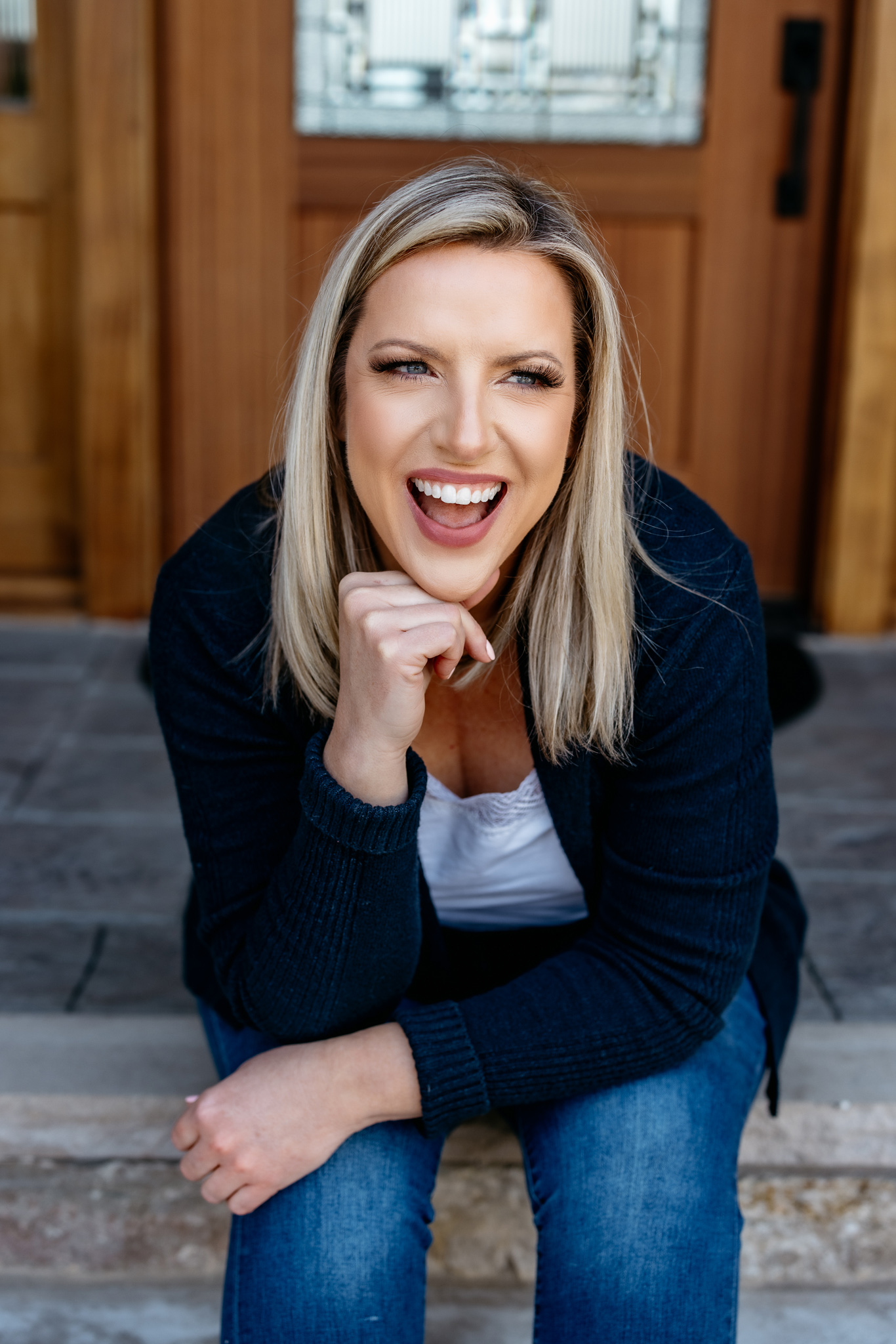 A blonde lady in a black sweater and blue jeans sitting on the steps in front of a brown door