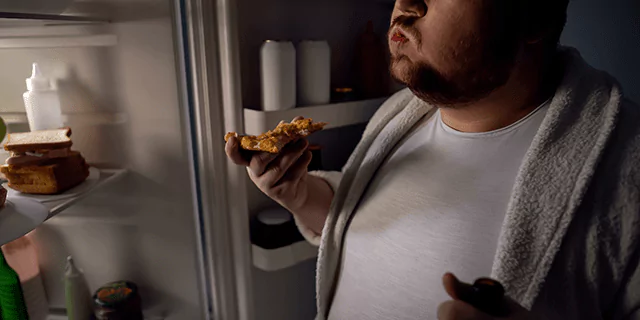 a person eating a slice of pizza near opened fridge at night