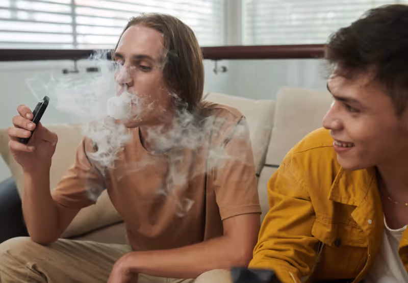 two boys sitting on a couch while using a vape