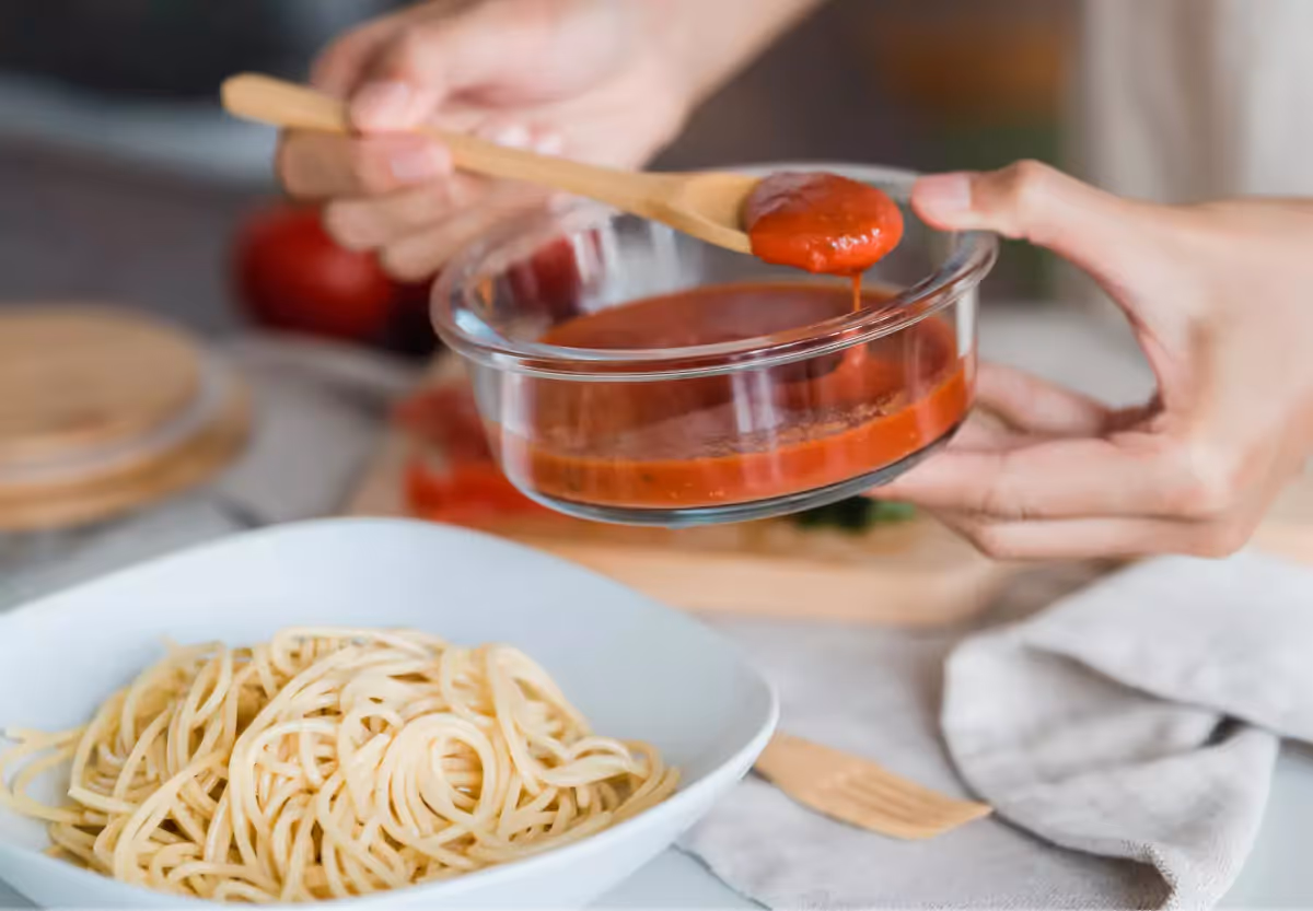 A closeup of hands holding a wooden spoon and a bowl of red pasta sauce