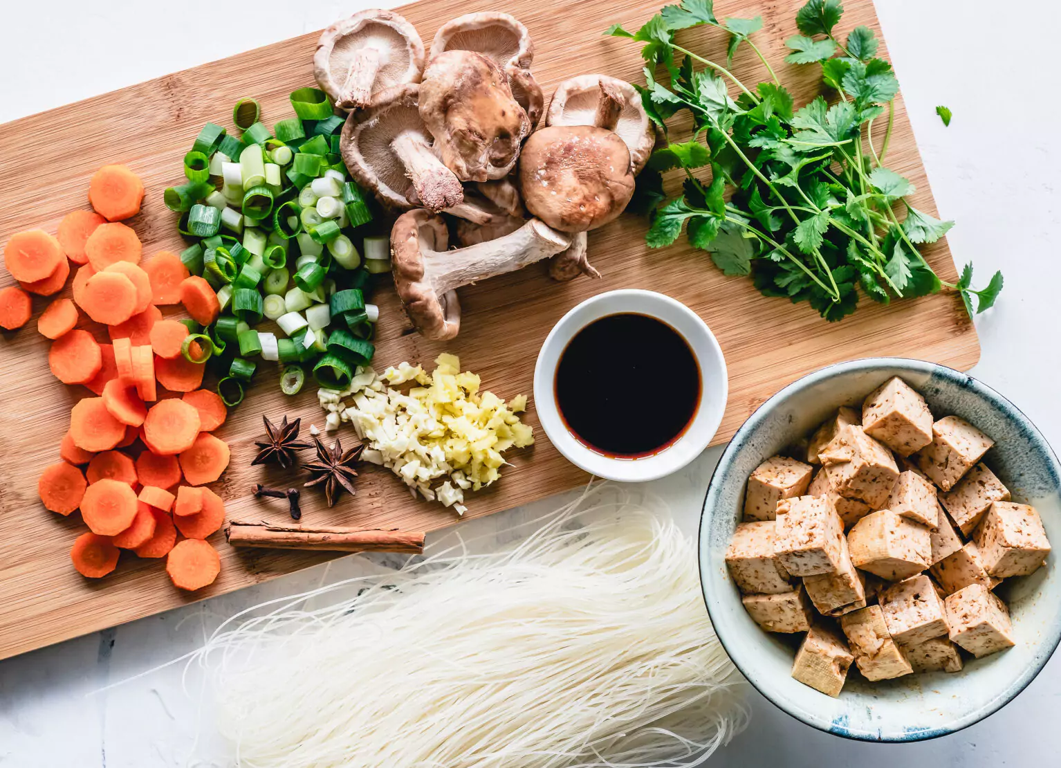 A chopping board with mushrooms, carrots, green onions, cilantro, garlic, cloves, cinnamon, soy sauce, tofu, and noodles
