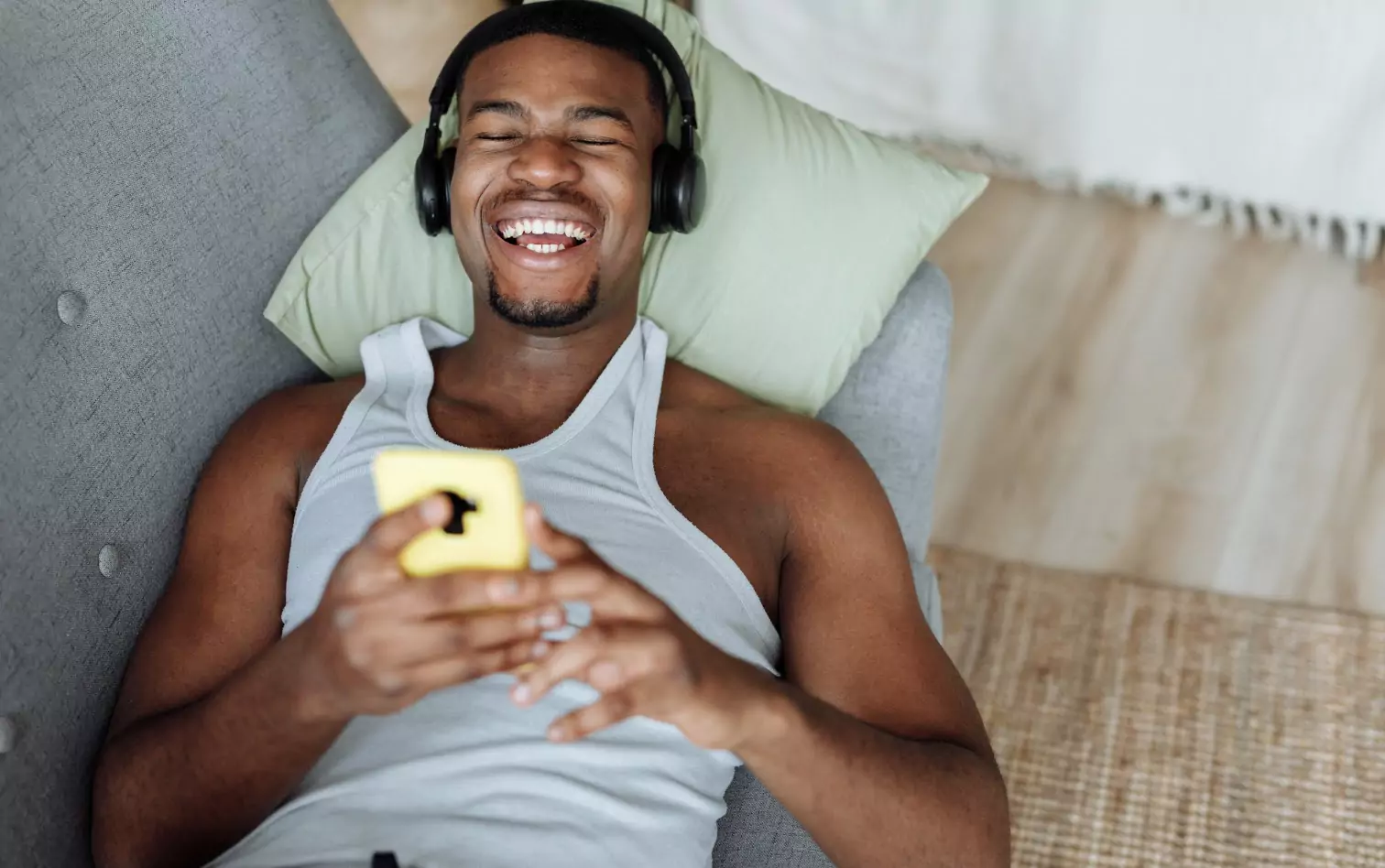 A man wearing headphones, holding a smartphone, lying on his back and laughing