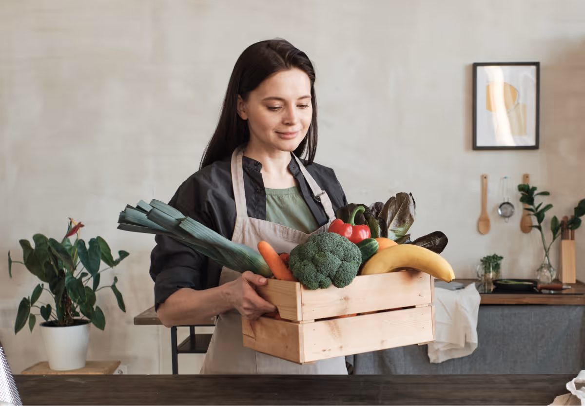 Someone with long dark hair, carrying a wood box of assorted fresh fruits and vegetables 
