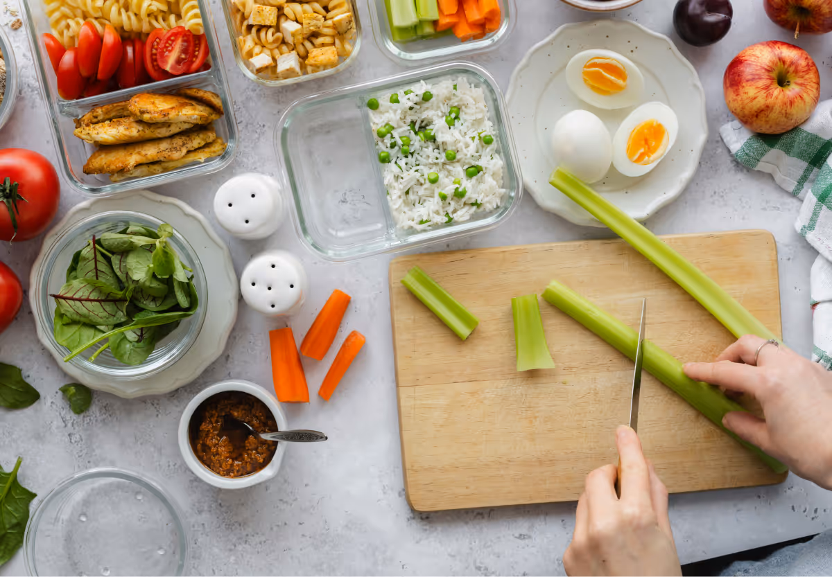 An array of food in glass storage boxes and white plates with a wooden chopping board and persons hands chopping celery in front of it