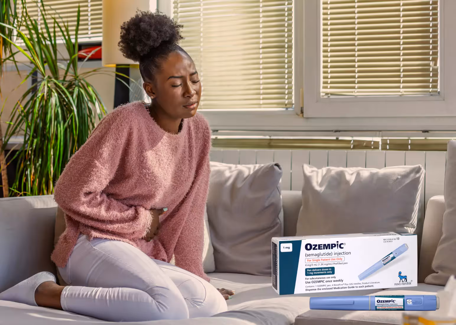 Someone with black hair tied in a bun, sitting on a couch in a pink sweater, holding their stomach. Box of ozempic overlaid